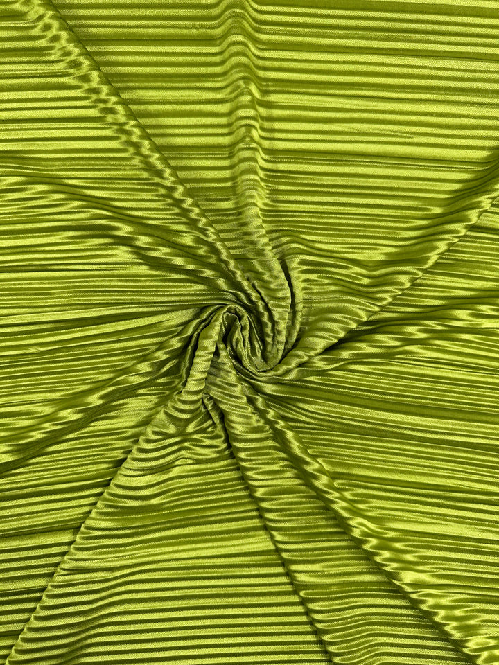 A detailed view of the chartreuse Metallix Pleat polyester fabric by Super Cheap Fabrics displays its pleated arrangement in a spiral pattern, offering a textured and wavy appearance. The lightweight folds capture the light, enhancing the shiny and smooth finish of this 140cm wide fabric.