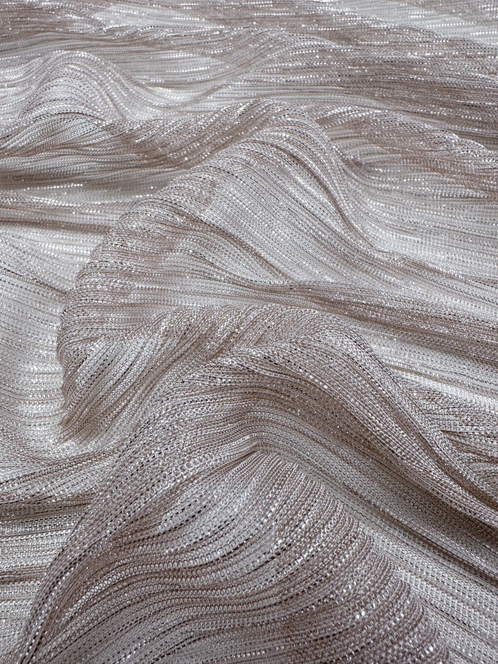 A close-up view of the Metallix Pleat in Blush by Super Cheap Fabrics showcases the lightweight, textured fabric adorned with a wavy pattern enhanced by metallic and shimmering threads. The polyester surface exudes a soft, flowing appearance, enriched with intricate cream, blush, and silver tones that beautifully capture and reflect light.