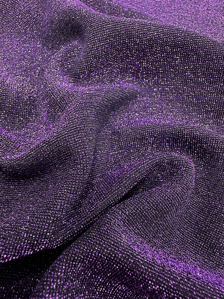 A close-up view of the Metallic Lycra - Disco Plum - 160cm by Super Cheap Fabrics shows its striking purple and black hues with a shimmering metallic finish. Ideal for crafting stylish dresses or tops, this fabric is draped gracefully, with its folds forming wavy patterns that accentuate its reflective qualities.