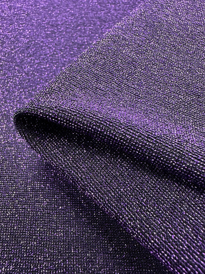 A close-up view of the shimmering Disco Plum Metallic Lycra from Super Cheap Fabrics reveals its luxurious purple hue and metallic texture. Folded to highlight its reflective quality and detailed weave, this fabric is perfect for dresses or tops, creating a dazzling effect in the light. It measures 160cm wide.