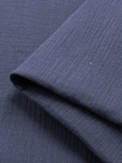 Close-up of folded navy muslin fabric from Super Cheap Fabrics, featuring a subtle textured pattern. The surface appears smooth with fine lines running across it, providing a slightly ribbed look.