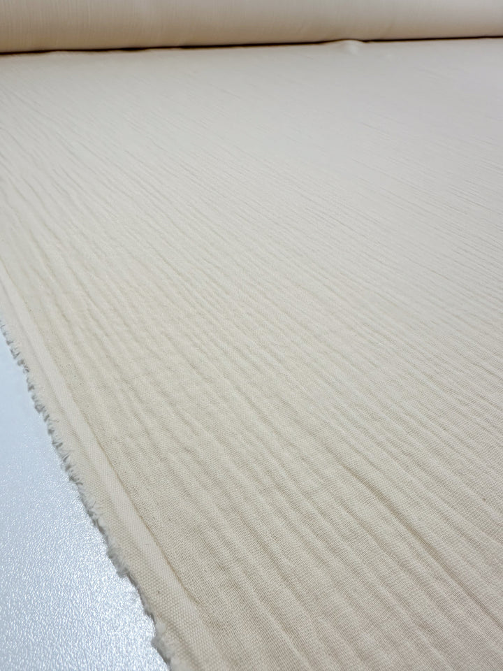 A large roll of Super Cheap Fabrics' Muslin - Almond Milk - 135cm is unrolled across a flat surface, showcasing its textured surface and selvage edge. This organic fabric appears soft, featuring subtle vertical lines, making it an ideal reusable material for various projects.