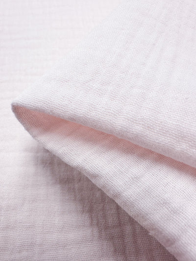 A close-up view showcases the Muslin - Slightly Pink - 135cm from Super Cheap Fabrics. The fabric features a soft, pale pink hue with a subtly textured, grid-like pattern. When folded, the reusable cloth highlights its lightweight and airy quality, and the visible texture suggests a gentle and delicate material.