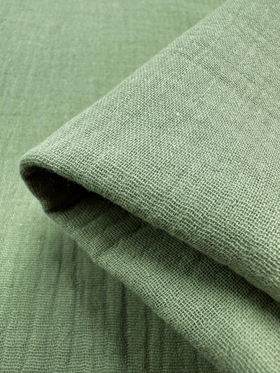 A detailed view of a folded piece of muslin fabric in laurel green from Super Cheap Fabrics reveals its woven pattern with subtle shading variations. This soft, lightweight fabric, reminiscent of organic linen, is perfect for reusable cloth projects.