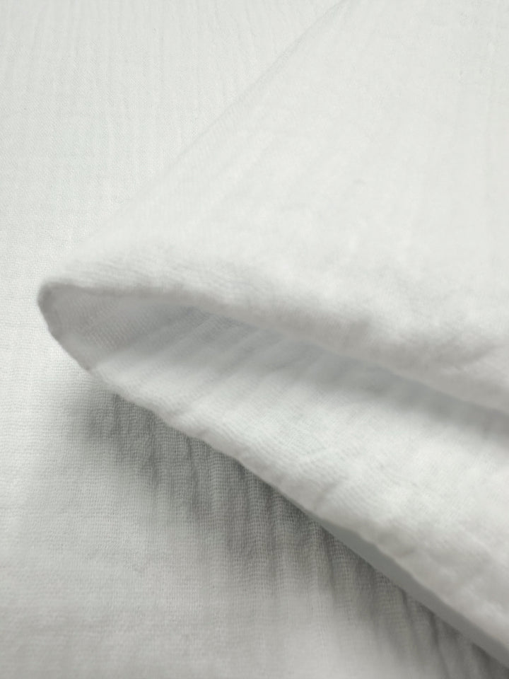 A close-up of the folded "Muslin - Snow White - 135cm" by Super Cheap Fabrics highlights its soft texture and lightweight quality. This organic fabric presents a smooth appearance with a subtle weave pattern, indicating it is breathable and comfortable, making it an excellent reusable material for various applications.