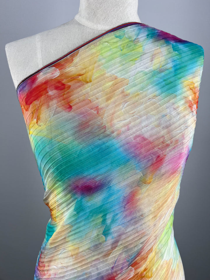 A mannequin is draped with *Textured Tencel - Candy Clouds - 136cm* from Super Cheap Fabrics, showcasing a pleated fabric in a watercolor pattern with vibrant shades of blue, orange, pink, yellow, and green. This sustainable material enhances its soft and artistic appearance.