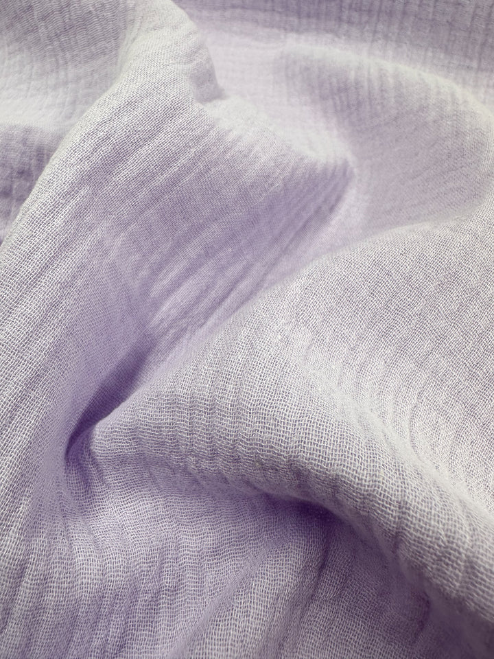 Close-up of a soft, textured light purple fabric labeled "Muslin - Lilac Breeze - 137cm" by Super Cheap Fabrics, featuring gentle folds and visible woven patterns that create a cozy and delicate appearance.