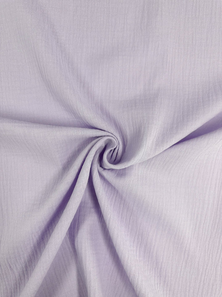 A close-up of Muslin - Lilac Breeze, an organic fabric from Super Cheap Fabrics, showcases a light purple hue with subtle lines and swirls, elegantly gathered into a spiral fold in the center.