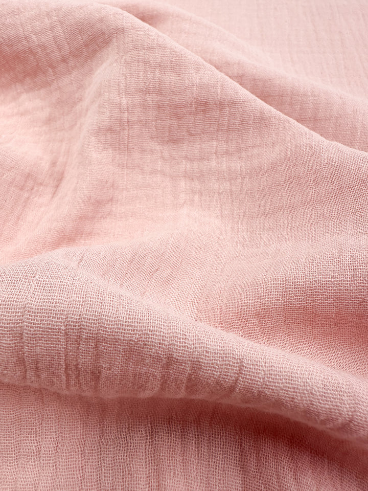 A close-up of the Super Cheap Fabrics' Muslin - Lotus - 135cm showcases its pink textured muslin with soft folds that create gentle shadows and highlights. The fabric is lightweight and finely woven, featuring a subtle grid pattern, ideal for those seeking organic elegance.