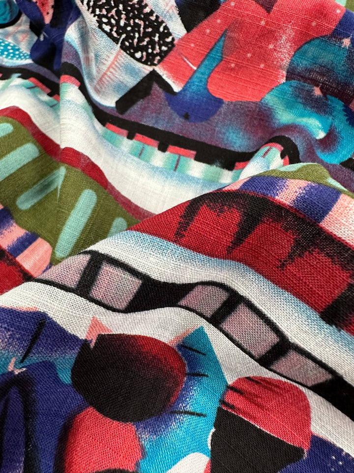 A detailed view of the Linen Blend - Mapalo - 145cm from Super Cheap Fabrics showcases its vibrant, abstract design with wavy folds. The geometric shapes in red, blue, black, white, and green create a dynamic and colorful texture on this breathable canvas.