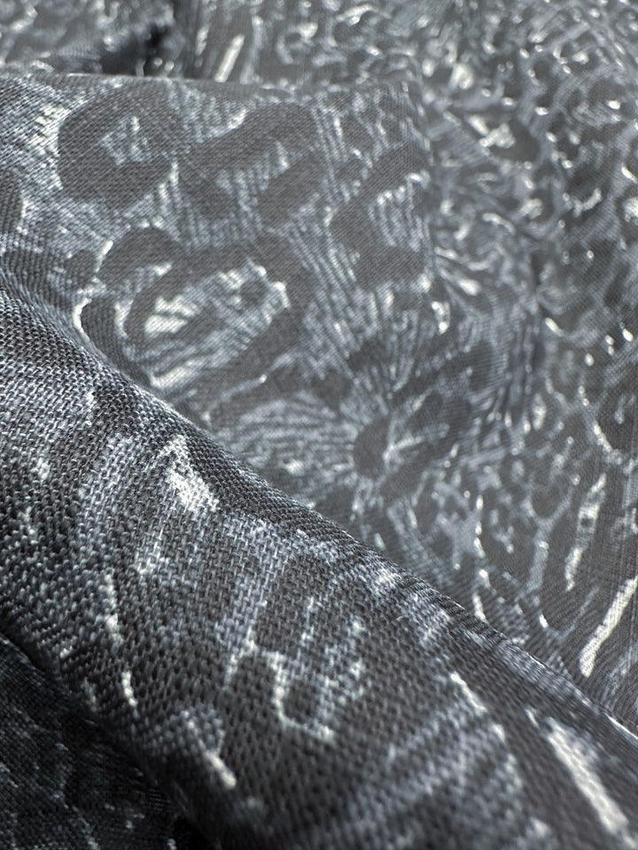 A close-up view of the "Linen Blend - Cool Jungle - 140cm" by Super Cheap Fabrics reveals its gray, lightweight linen fabric featuring a shiny, textured surface with an abstract pattern. The way the light reflects off the raised areas produces a dynamic visual effect, making it ideal for summer outfits.