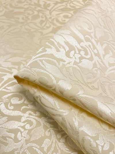 The Designer Damask - Rutabaga - 155cm fabric from Super Cheap Fabrics features an intricate, swirling floral pattern on cream-colored jacquard, reminiscent of luxurious damask. Its subtle sheen beautifully highlights the embossed design.