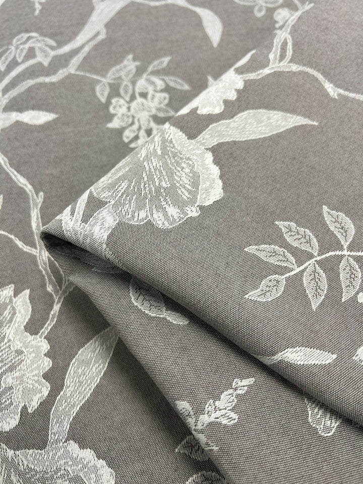 Close-up of folded Designer Damask - Saphire Dust - 140cm fabric by Super Cheap Fabrics set against a gray backdrop, highlighting an intricate white floral pattern. The luxurious fabric showcases detailed leaves and flowers, exuding an elegant and sophisticated design.