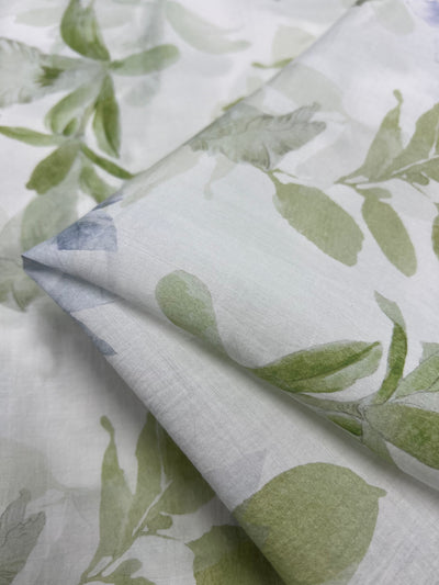 Close-up of the Designer Cotton - Lotus - 150cm from Super Cheap Fabrics, featuring a subtle, nature-inspired pattern with green leaves. The fabric is folded, showcasing its texture and soft botanical design—perfect for luxury sewing projects.