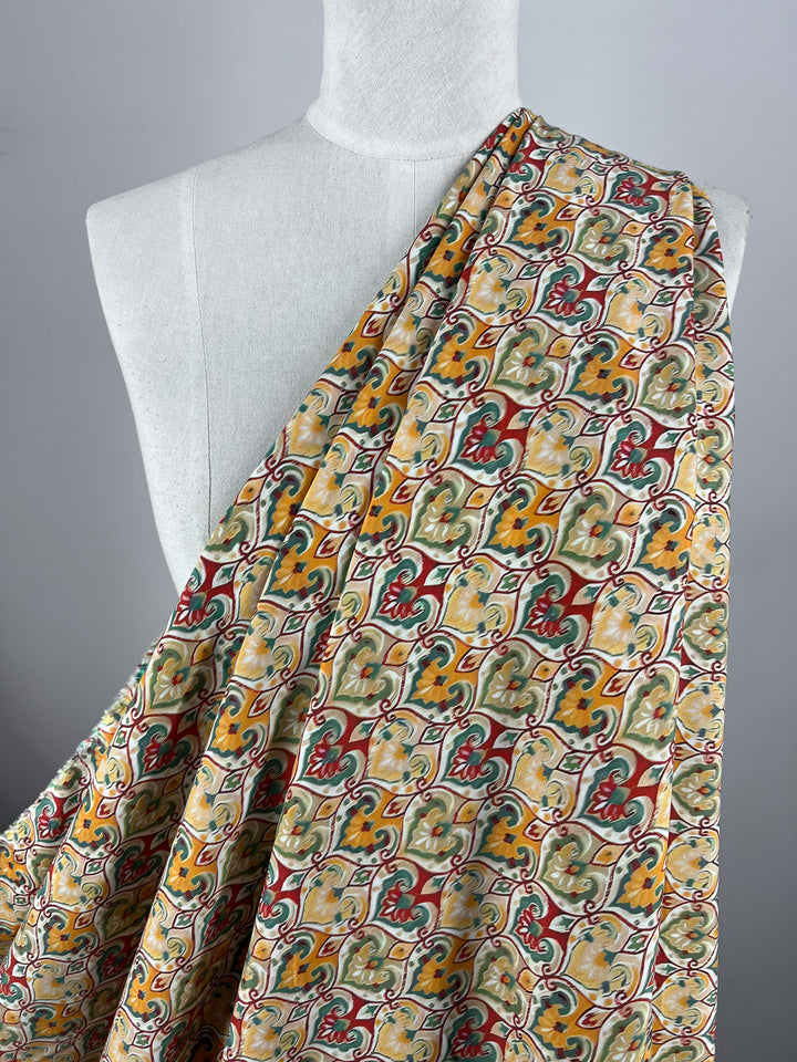 A mannequin draped in the Designer Rayon - Belle - 145cm by Super Cheap Fabrics showcases a colorful, intricate floral and geometric pattern in red, green, yellow, and beige on luxurious rayon fabric. The simple gray background highlights the detailed design, making it a versatile choice for any fashion enthusiast.