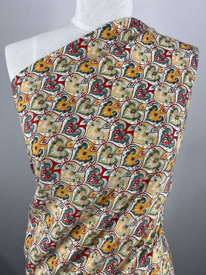 A mannequin draped in the Designer Rayon - Belle - 145cm from Super Cheap Fabrics showcases a colorful, intricate floral and geometric pattern in shades of yellow, red, green, and white. The versatile material is elegantly wrapped around the shoulder, highlighting its stunning design.