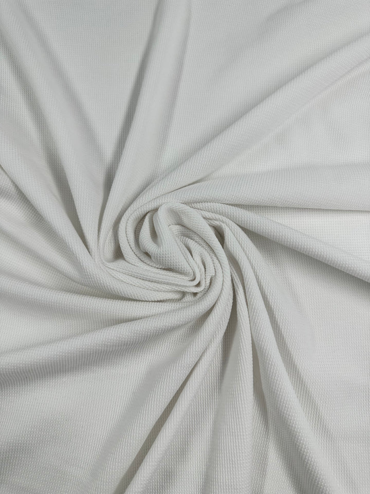 Close-up of a white Honeycomb Knit - White - 158cm fabric by Super Cheap Fabrics twisted in a spiral pattern. The fabric appears soft and has a subtle grid-like weave, creating small squares across its surface. The twist in the fabric forms a gentle, flowing spiral in the center, showcasing its unique texture.
