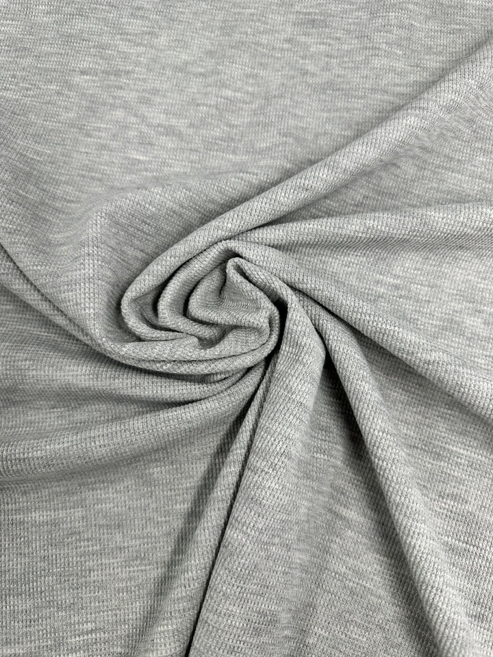 Close-up of a light gray honeycomb fabric with a textured surface, artfully gathered and twisted in the center, creating soft folds and a swirling pattern. The material appears stretchy and comfortable, typical of jersey or knit fabric. The Honeycomb Knit - Light Grey - 158cm by Super Cheap Fabrics exemplifies these qualities vividly.