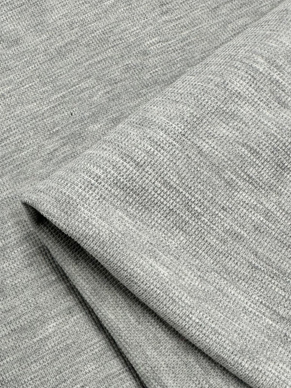 Close-up view of a grey fabric with a soft, textured surface. The fabric appears to be folded, creating overlapping layers and showcasing a subtle, fine-knit pattern. This detail gives the appearance of a smooth yet slightly heathered finish with a hint of three-dimensional effect. The product is Honeycomb Knit - Light Grey - 158cm by Super Cheap Fabrics.