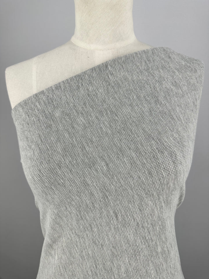 The image shows the upper part of a mannequin wearing a light gray, one-shoulder sweater made of fine, knitted fabric with a subtle three-dimensional effect. The background is plain and neutral. The fabric used is Honeycomb Knit - Light Grey - 158cm by Super Cheap Fabrics.