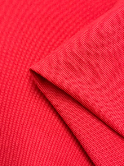 Close-up of a piece of bright red Honeycomb Knit - Watermelon - 158cm fabric by Super Cheap Fabrics with a woven texture. The fabric is folded diagonally, creating a visible crease and highlighting the smooth, uniform texture and vibrant color, adding a subtle three-dimensional effect.