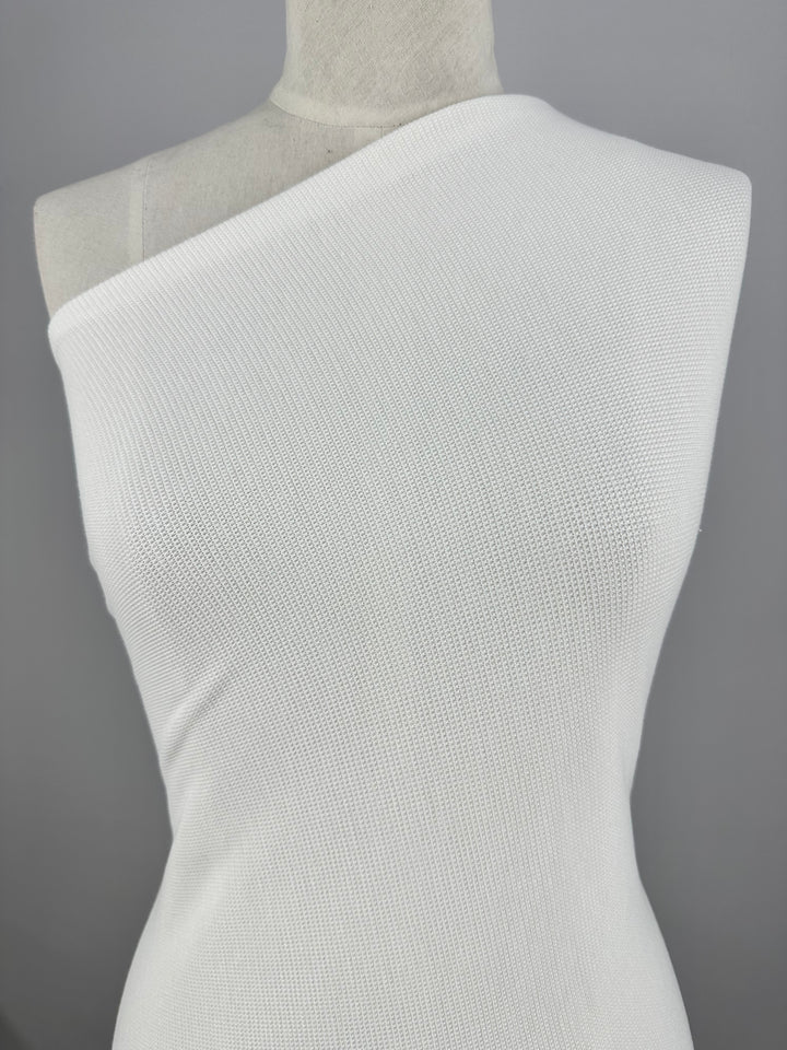 A white Waffle Knit - White - 152cm from Super Cheap Fabrics displayed on a mannequin against a grey background. The fabric, resembling honeycomb fabric, has a subtle ribbed texture, giving it a slightly textured and three-dimensional effect. The headless mannequin focuses attention on the dress's sleek and minimalist design.