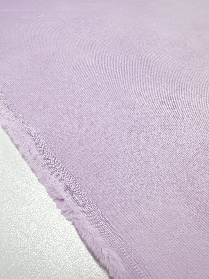 A close-up of the Super Cheap Fabrics' Micro Wale Corduroy - Lavender Frost shows its ribbed texture and fringed edges against a smooth white surface.