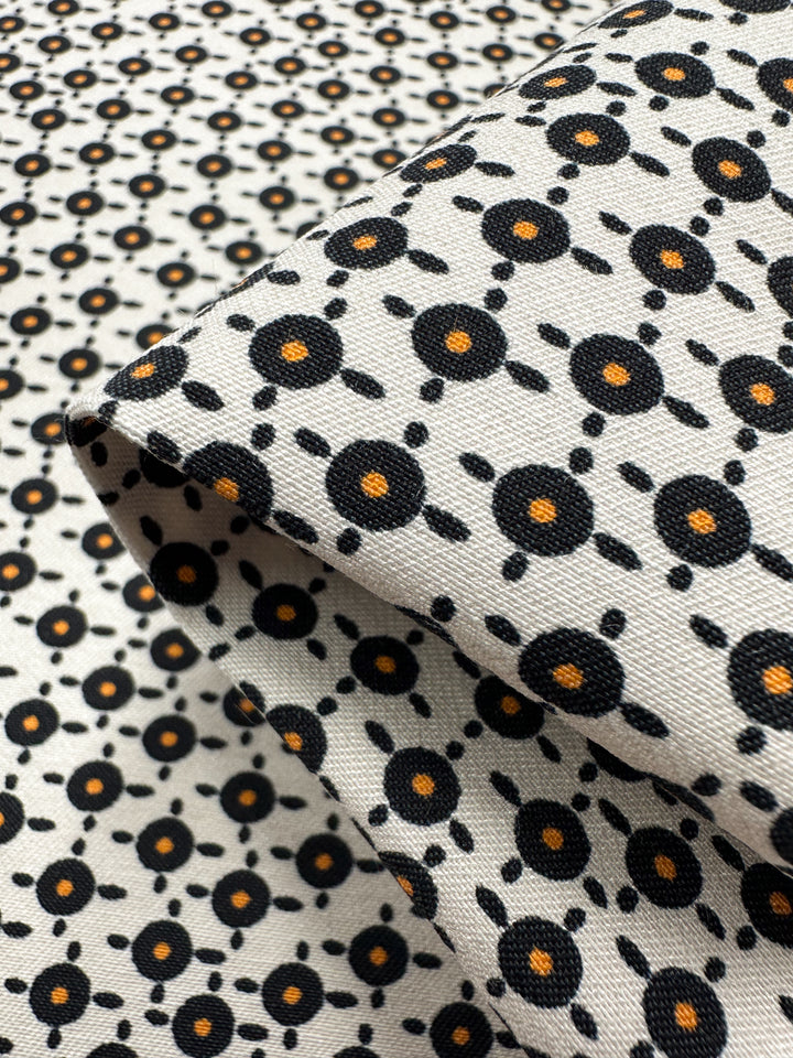 The fabric displays a pattern of black circles with small orange dots, connected by a grid on a white background. The medium-weight stretch is folded to reveal its repetitive design. Product: Printed Bengaline - Radar - 150cm by Super Cheap Fabrics.