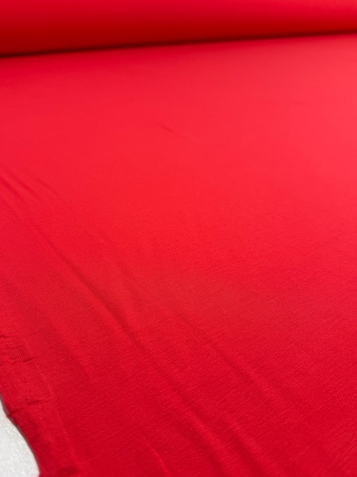 A close-up of Super Cheap Fabrics' Cotton Lycra - Hibiscus - 165cm shows the smooth, bright red fabric laid flat with subtle folds and wrinkles, its stretchy surface extending from a rolled edge at the top.
