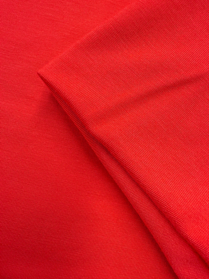A close-up of Super Cheap Fabrics' Cotton Lycra - Hibiscus (165cm) shows the smooth, bright red stretchy fabric with a folded corner, highlighting its texture and weave.