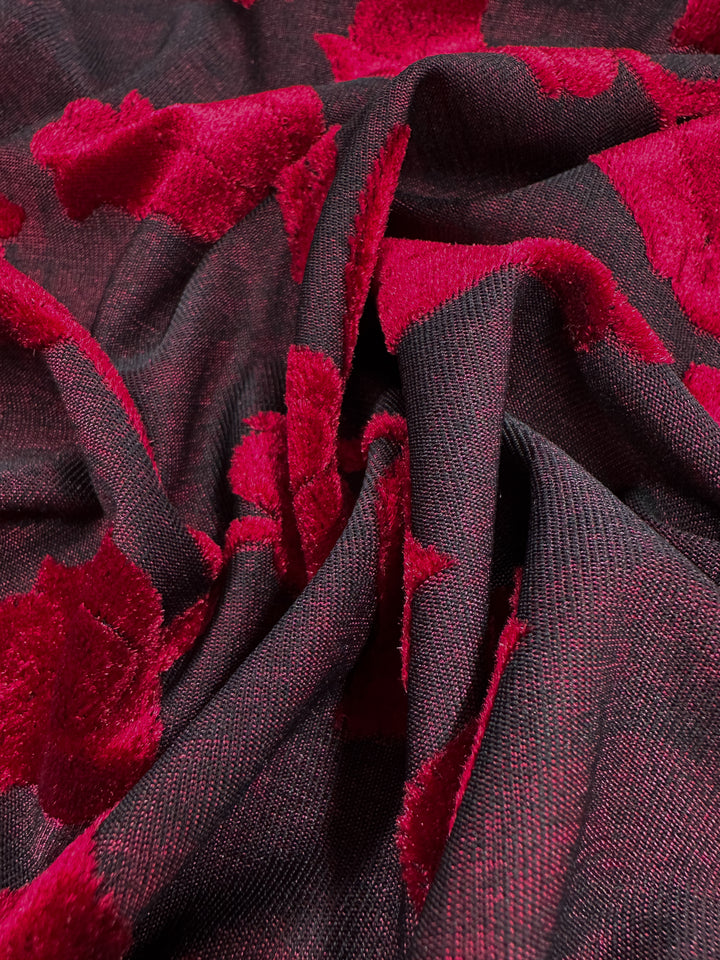 A close-up showcases the luxurious Burnout Velvet - Lychee from Super Cheap Fabrics. It features a dark base with rich red floral patterns in an intricate burnout design, creating a dramatic contrast that highlights the exquisite details and folds of the material. Width: 150cm.