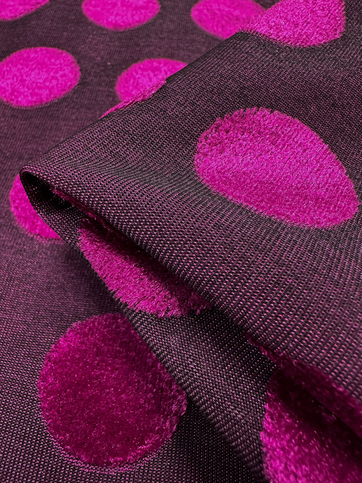 The Burnout Velvet - Festival Fuchsia fabric from Super Cheap Fabrics features a dark base with large, textured bright pink polka dots. The fabric folds highlight the contrast between its smooth background and velvety dots, showcasing exquisite textile artistry.