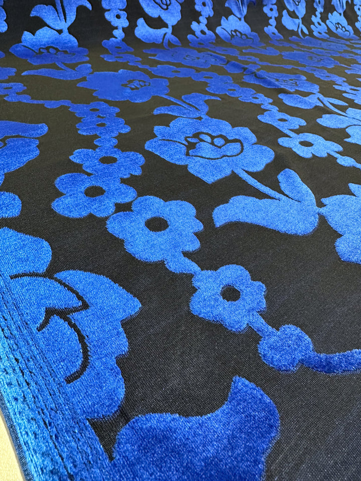 A close-up reveals Super Cheap Fabrics' Burnout Velvet in Snorkel Blue (155cm), showcasing a bold floral pattern in vibrant blue velvet on black, with assorted floral shapes and stems for an elegant, textured look.