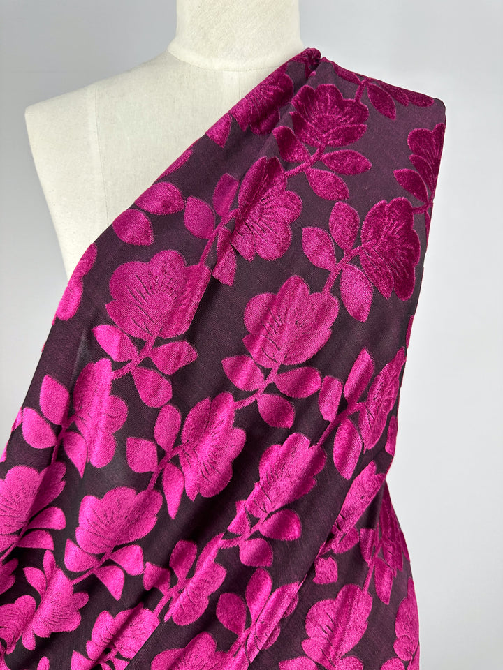 A mannequin showcases Super Cheap Fabrics' Burnout Velvet in Cactus Flower - 160cm, with a luxurious dark purple shade and vibrant pink floral pattern. It drapes elegantly over one shoulder, highlighting its intricate design against a plain background.