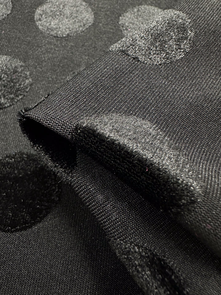 The Burnout Velvet - Polka Black by Super Cheap Fabrics features luxurious, large shiny polka dots on a matte background. The striking visual effect is enhanced when the 155cm-wide fabric folds over itself, accentuating the contrast between its textures.