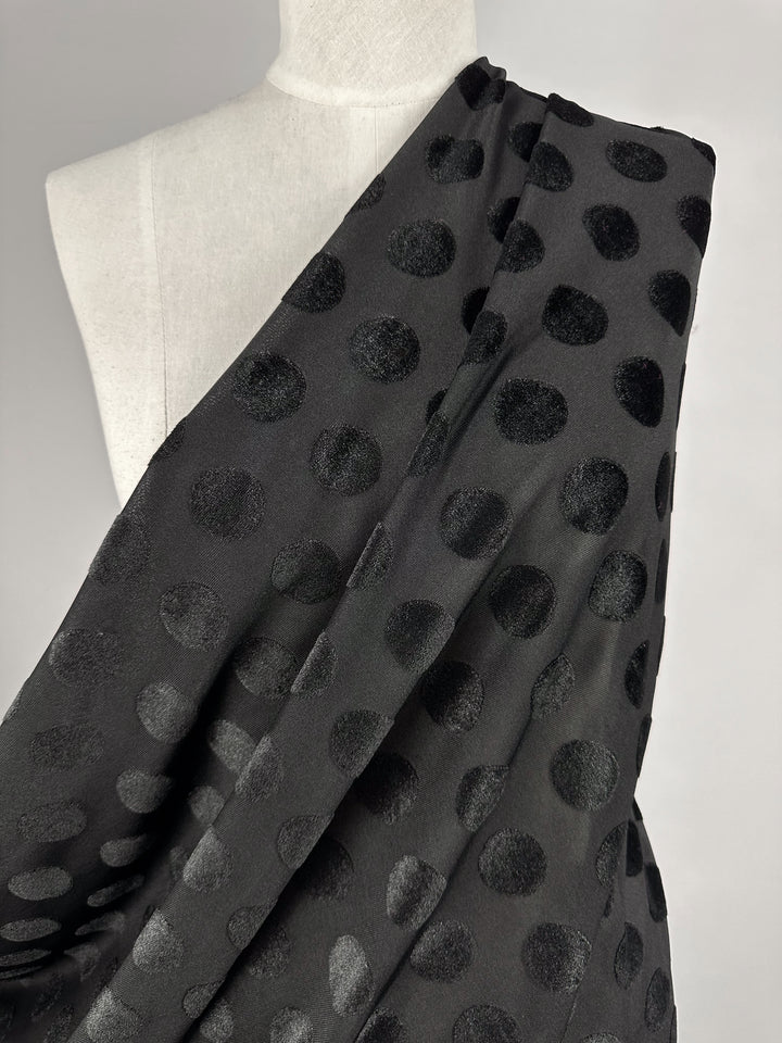 A close-up of Burnout Velvet - Polka Black by Super Cheap Fabrics, featuring glossy polka dots on plush black velvet. Elegantly draped over a mannequin, it creates a striking visual against a plain backdrop.