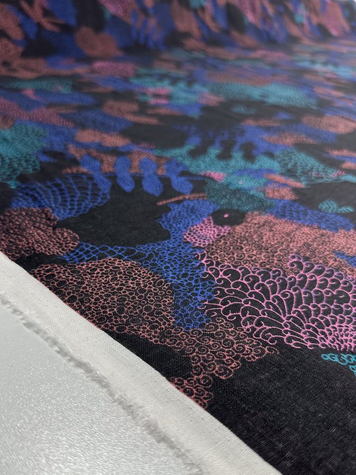 Close-up of the Linen Blend - Coral - 147cm by Super Cheap Fabrics, showcasing a lightweight fabric with an intricate abstract design in pink, blue, and purple on a multi black background. One edge appears unfinished with a visible white selvage.