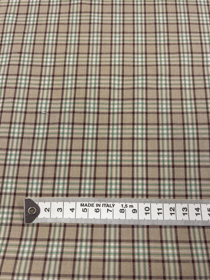 Image depicting a close-up of the Shirting - Safari - 148cm fabric by Super Cheap Fabrics, featuring a light to medium weight checkered pattern in green and brown set against a beige background. A white measuring tape marked "Made in Italy" runs along the bottom edge, showing measurements up to 13 centimeters. This polyester cotton fabric is ideal for crafting children's wear.