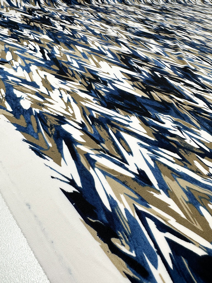 A close-up of Super Cheap Fabrics' Printed Lycra - Arrows (150cm), a medium-weight polyester/spandex fabric in blue, brown, and white with an abstract zigzag design covering the entire surface.