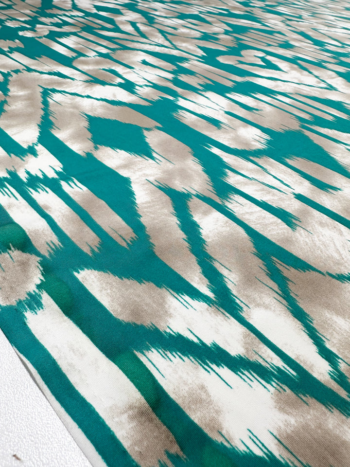 A close-up shows the medium-weight Printed Lycra - Dynasty - 150cm from Super Cheap Fabrics, with a turquoise and cream abstract pattern. Crafted from Polyester/Spandex, the jagged brush stroke design creates a dynamic look as it lies flat on a surface.