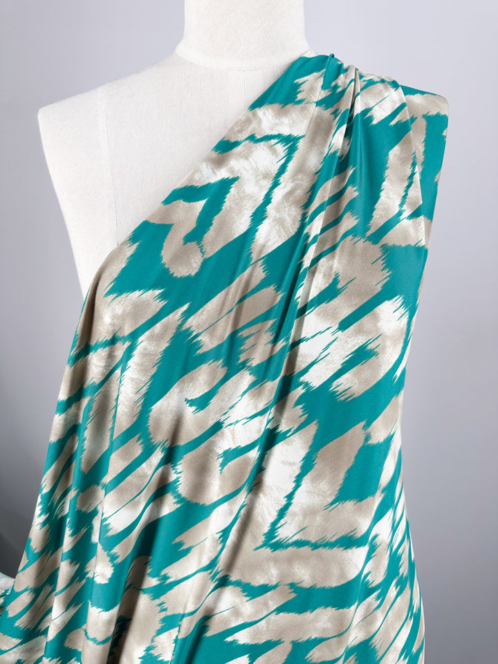 A mannequin draped in Super Cheap Fabrics' Printed Lycra - Dynasty - 150cm features a bold turquoise and beige zigzag pattern. The medium-weight Polyester/Spandex fabric elegantly covers one shoulder, showcasing the dynamic design.