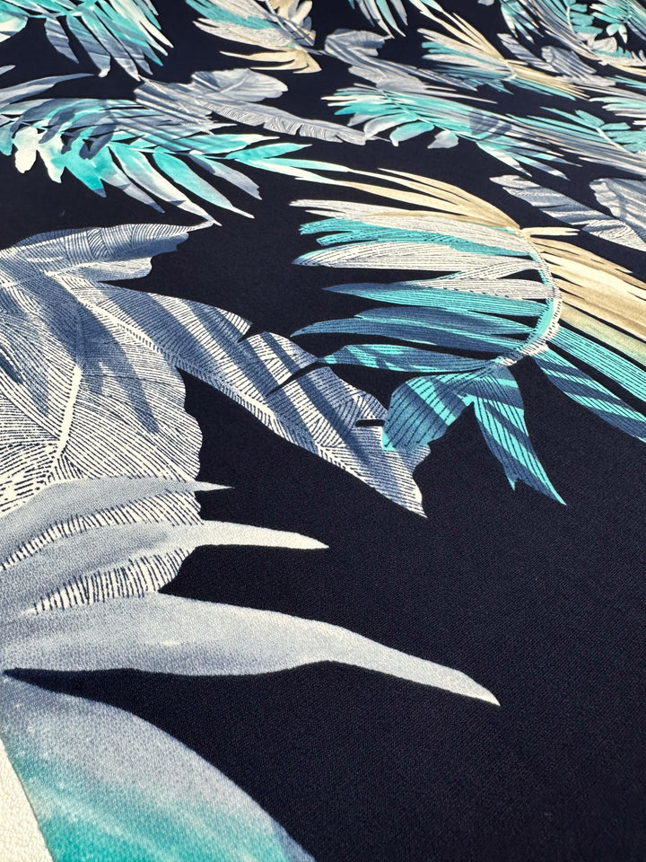 Super Cheap Fabrics' Printed Lycra - Frost Tropic - 150cm features medium-weight fabric with a blue, teal, and white tropical leaf pattern on a dark background. The overlapping palm leaves create a vibrant layered look, offering added stretch and comfort.