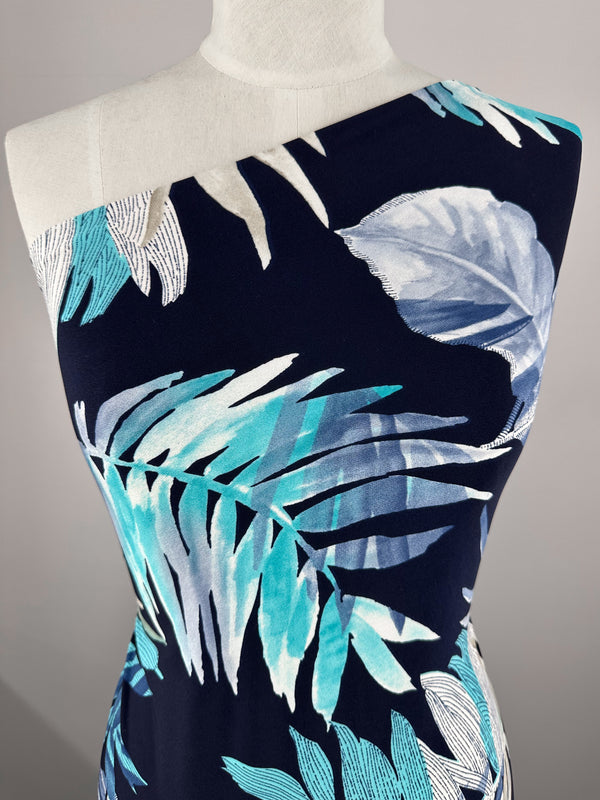 A mannequin displays a one-shoulder dress featuring a bold, tropical leaf print in blue, white, and teal on a dark base. Made from Super Cheap Fabrics' "Printed Lycra - Frost Tropic - 150cm," it combines comfort and durability with medium-weight Polyester/Spandex material.