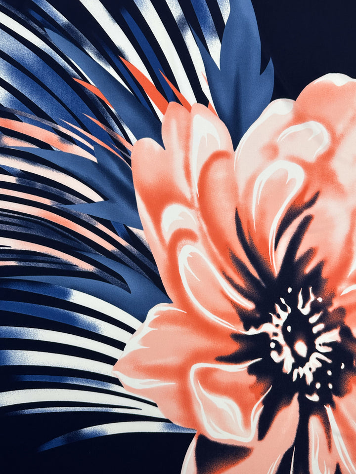 This printed Lycra from Super Cheap Fabrics features a vivid coral-colored flower with white and dark centers, set against blue and white abstract leaves on a dark background, creating dynamic contrast. It is a medium-weight fabric named "Printed Lycra - Bold - 150cm.