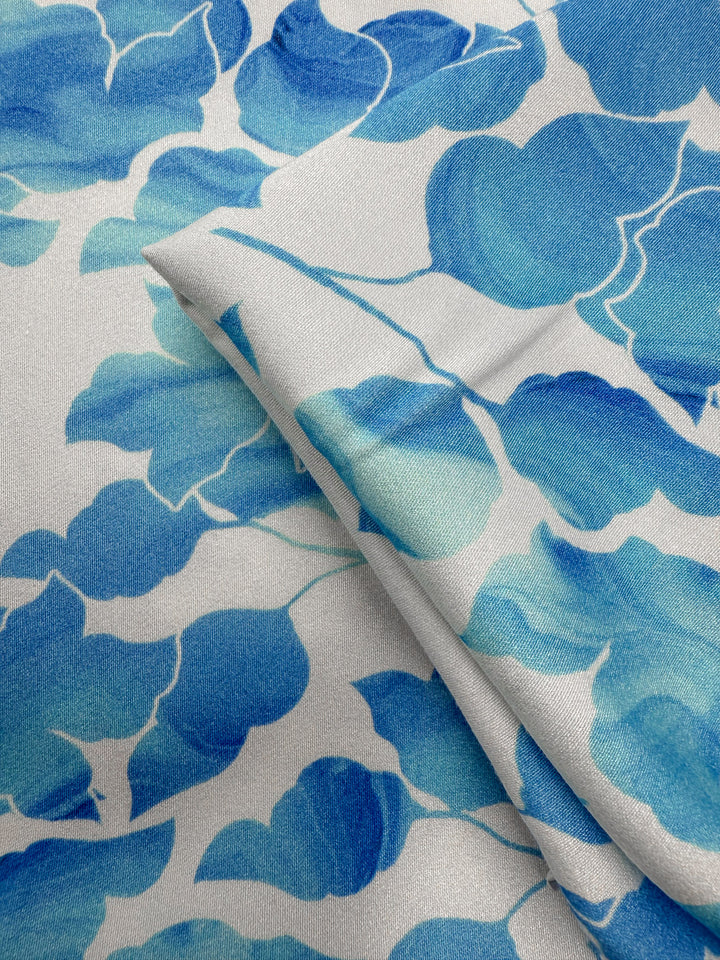 A close-up of Super Cheap Fabrics’ Mercerised Rayon - Blue Ginkgo (150cm), showing its lightweight feel, soft texture, and vibrant blue ginkgo leaf pattern on a light background, with one piece folded over another.