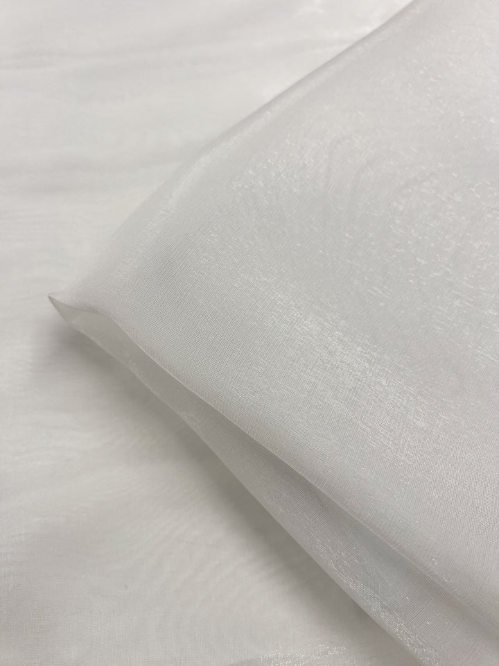 A close-up of the 'Silk Mousseline - Bone - 135cm' from Super Cheap Fabrics showcases its white, sheer silk fabric with an extra lightweight and a subtle metallic shimmer, neatly folded against a similar background. The texture appears smooth and delicate, exuding a soft, iridescent quality.