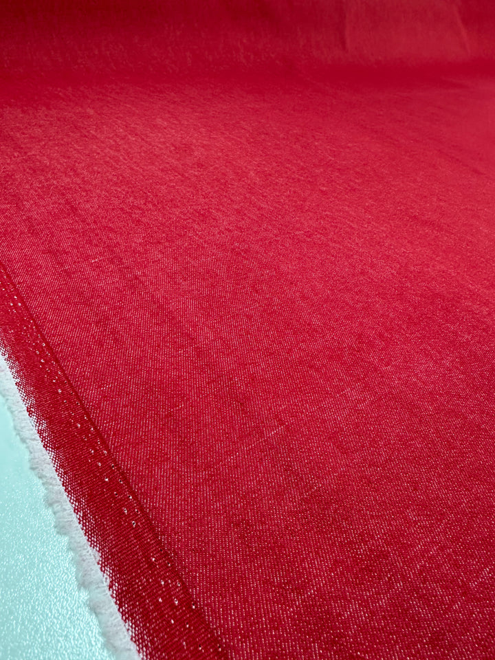 A close-up of Super Cheap Fabrics' Stretch Denim in Toreador red, 145cm wide. The fabric is medium weight with a smooth, slightly textured surface. The edge appears at the lower left corner, showing a subtle color variation under natural light.