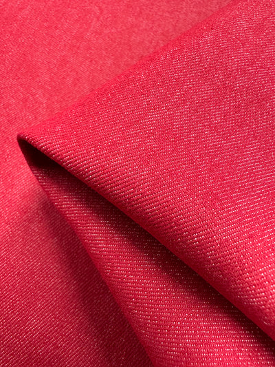 A close-up displays the folded Stretch Denim - Toreador - 145cm from Super Cheap Fabrics, showcasing the vibrant red color, subtle texture, and fine weave reminiscent of high-quality denim. The arrangement highlights its rich hue and detailed pattern.