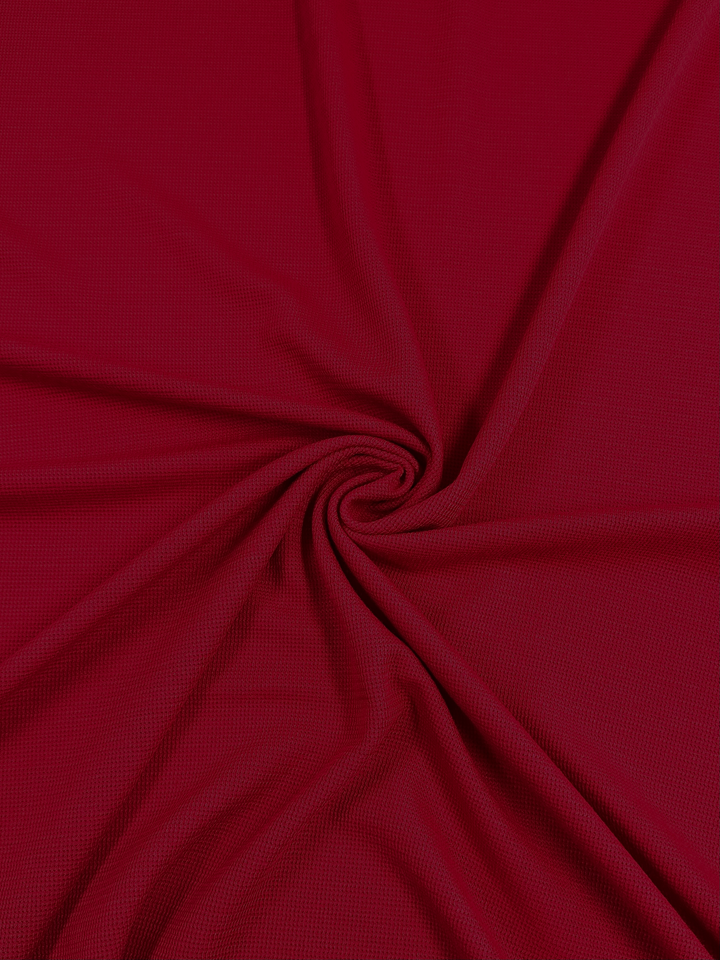 A piece of deep red Super Cheap Fabrics Waffle Knit - Fire - 152cm fabric is gathered and twisted into a circular pattern in the center. The smooth, slightly shiny texture interplays with a subtle three-dimensional effect, creating gentle folds and a swirled design.