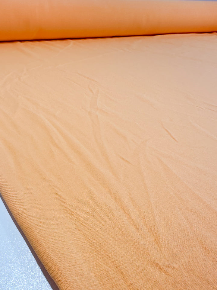 A close-up of the Super Cheap Fabrics' Cotton Jersey - Papaya, 160cm wide, shows the smooth medium weight cotton fabric in light papaya hue with slight wrinkles and folds on a flat surface.